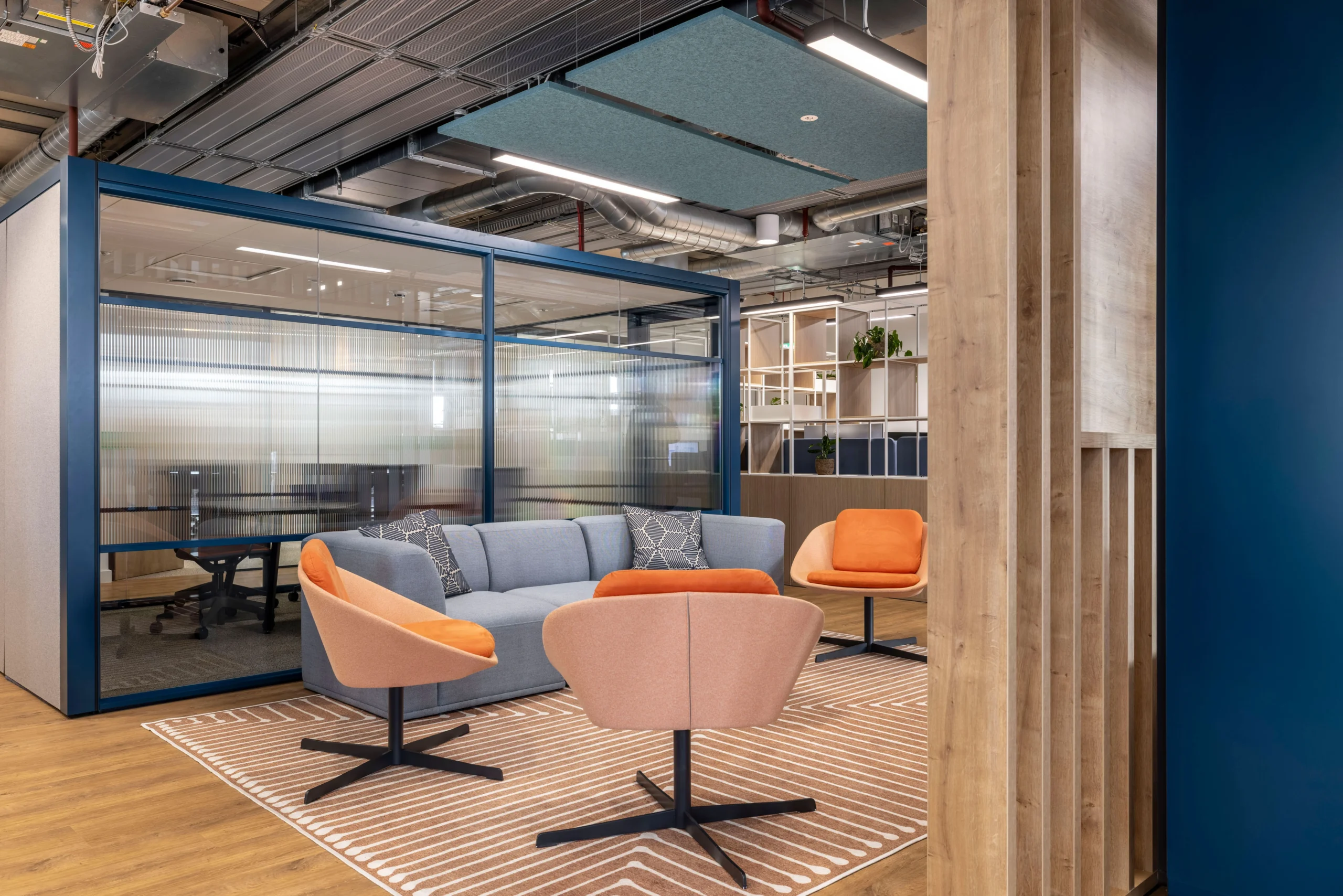 A collaborative workspace at Carbon Clean's office, designed by Modus Workspace, featuring a cosy seating arrangement with a grey sofa and orange chairs on a patterned rug. The space is enclosed by glass partitions for privacy while maintaining an open feel.