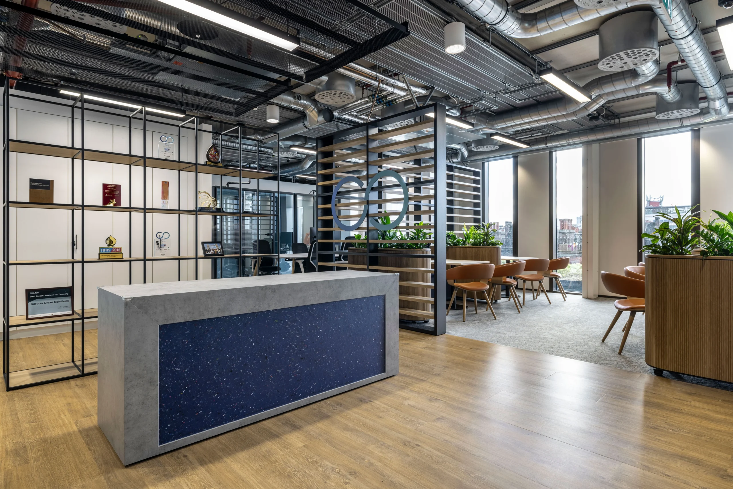 A reception area in Carbon Clean's office, designed by Modus Workspace, displaying a concrete reception desk and a stylish shelving unit with awards and decor. The area is surrounded by greenery and modern furnishings, providing a welcoming atmosphere.