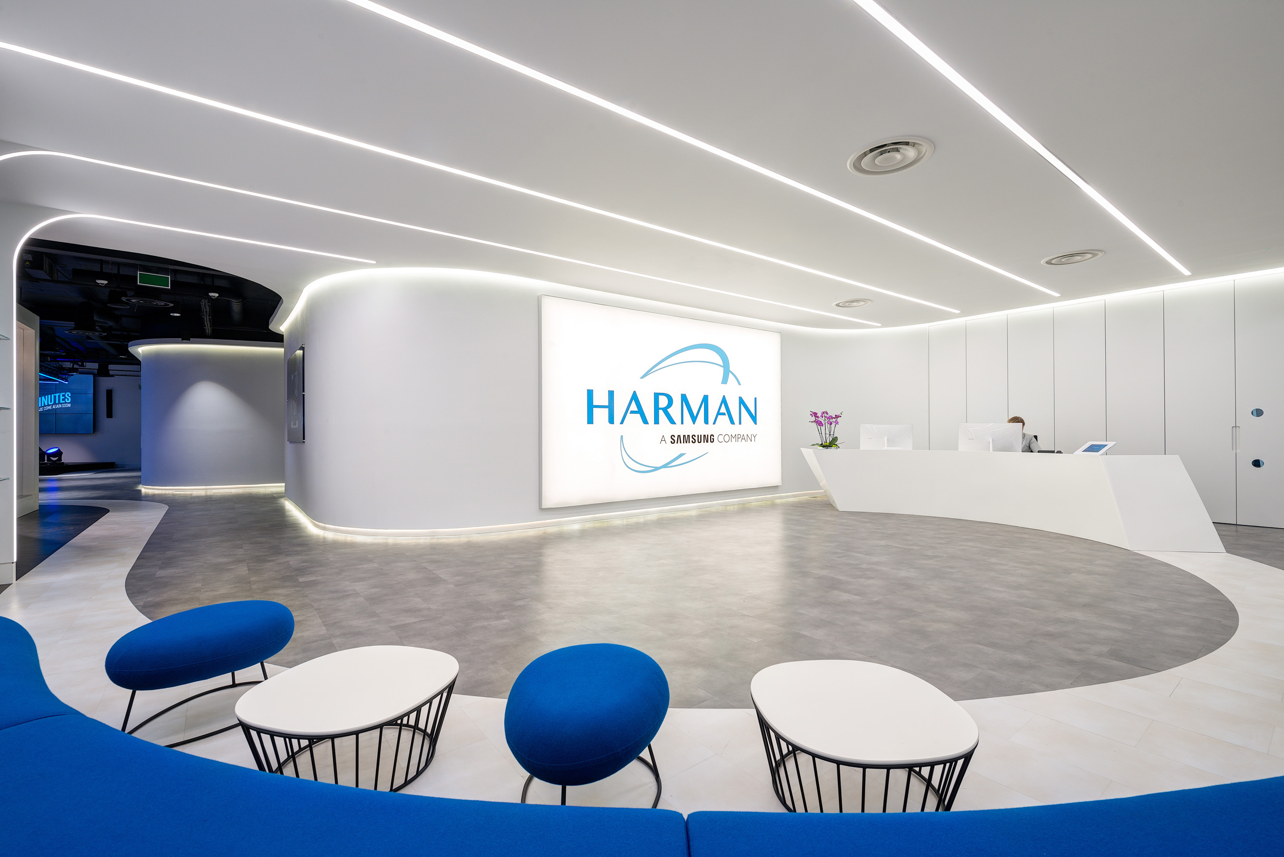 A modern reception area at Harman’s office features curved white walls, bright lighting strips, and a large, illuminated Harman logo. The area includes contemporary blue and white seating with round tables against grey and white flooring.