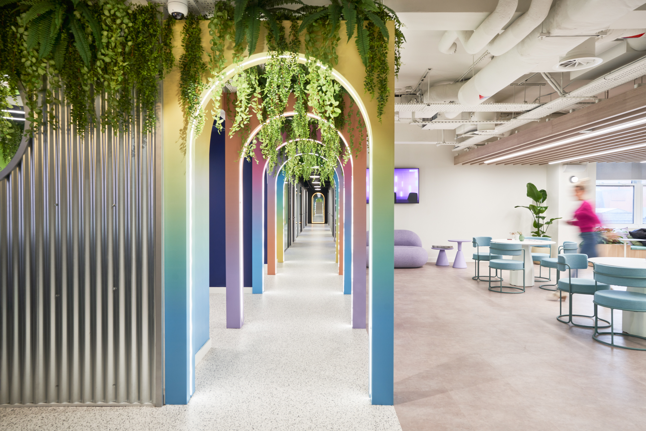 A colourful hallway at Huckletree, lined with illuminated rainbow-toned archways and hanging greenery, leading to an open communal area with modern seating and décor.