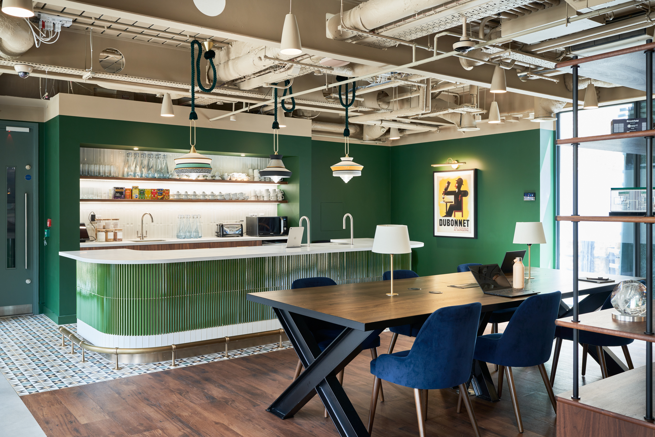 A modern and inviting teapoint and communal area designed by Modus Workspace for a private equity firm in Marylebone. The green tiled kitchen and blue velvet seating add elegance, while a framed vintage poster completes the ambiance.