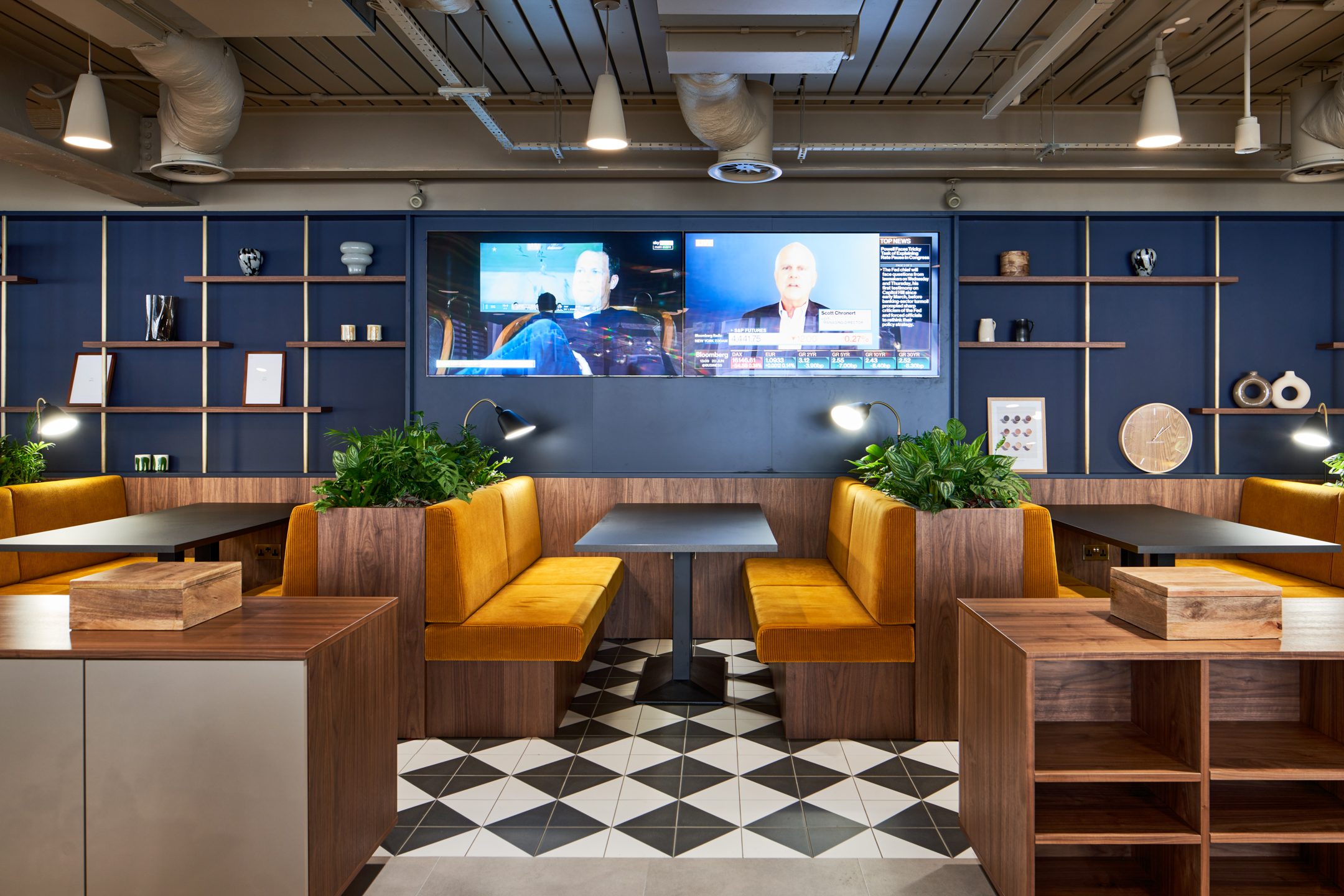 A cosy and vibrant booth seating area at a private equity firm in Marylebone, designed by Modus Workspace. The warm yellow seating paired with dark wood and geometric floor tiles offers a perfect space for casual meetings, with wall-mounted TVs in the background for presentations