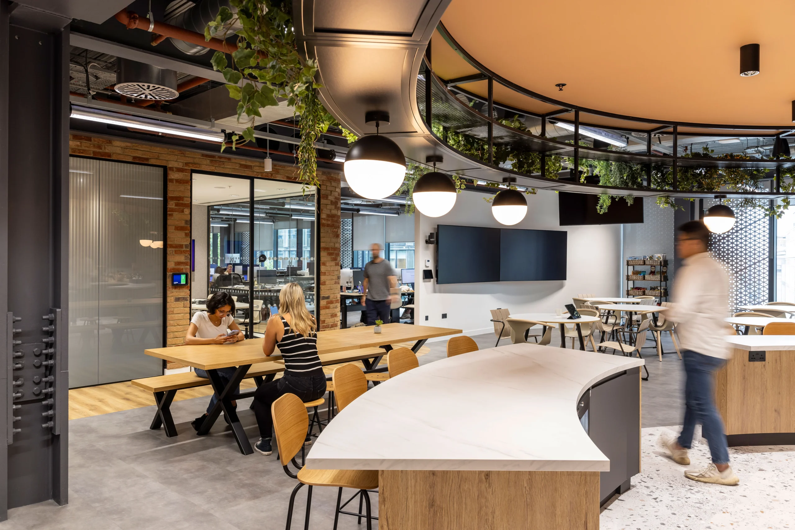 An open-plan workspace at Bazaarvoice's London office, designed by Modus Workspace, where employees are engaged in collaborative work. The area is filled with natural light, lush greenery, and modern decor, creating a dynamic and productive atmosphere that encourages teamwork and innovation.