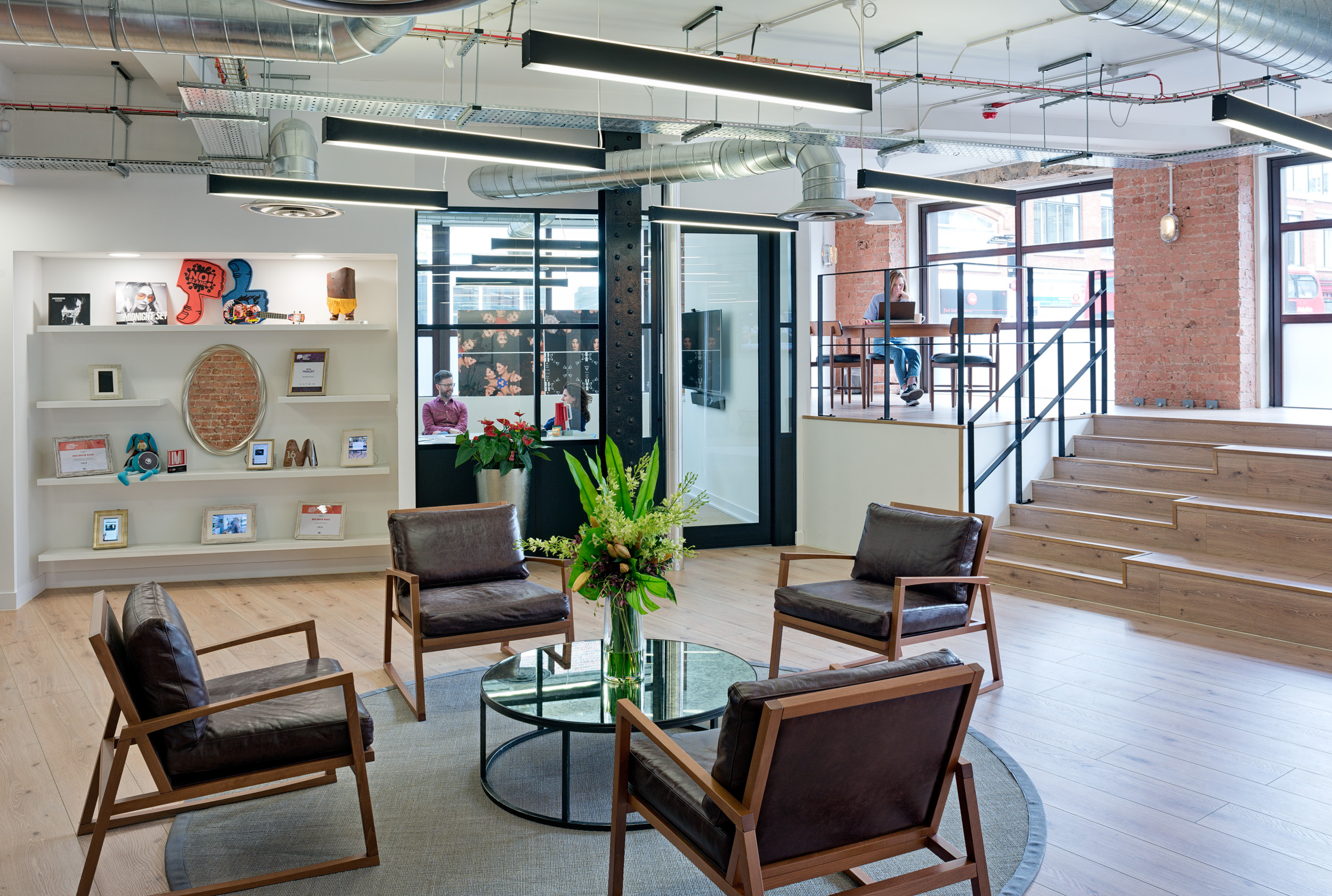 The reception waiting area in the Red Brick Road office features a modern industrial design with exposed brick and sleek furnishings. A timber staircase and bleacher setting lead to a mezzanine working station.