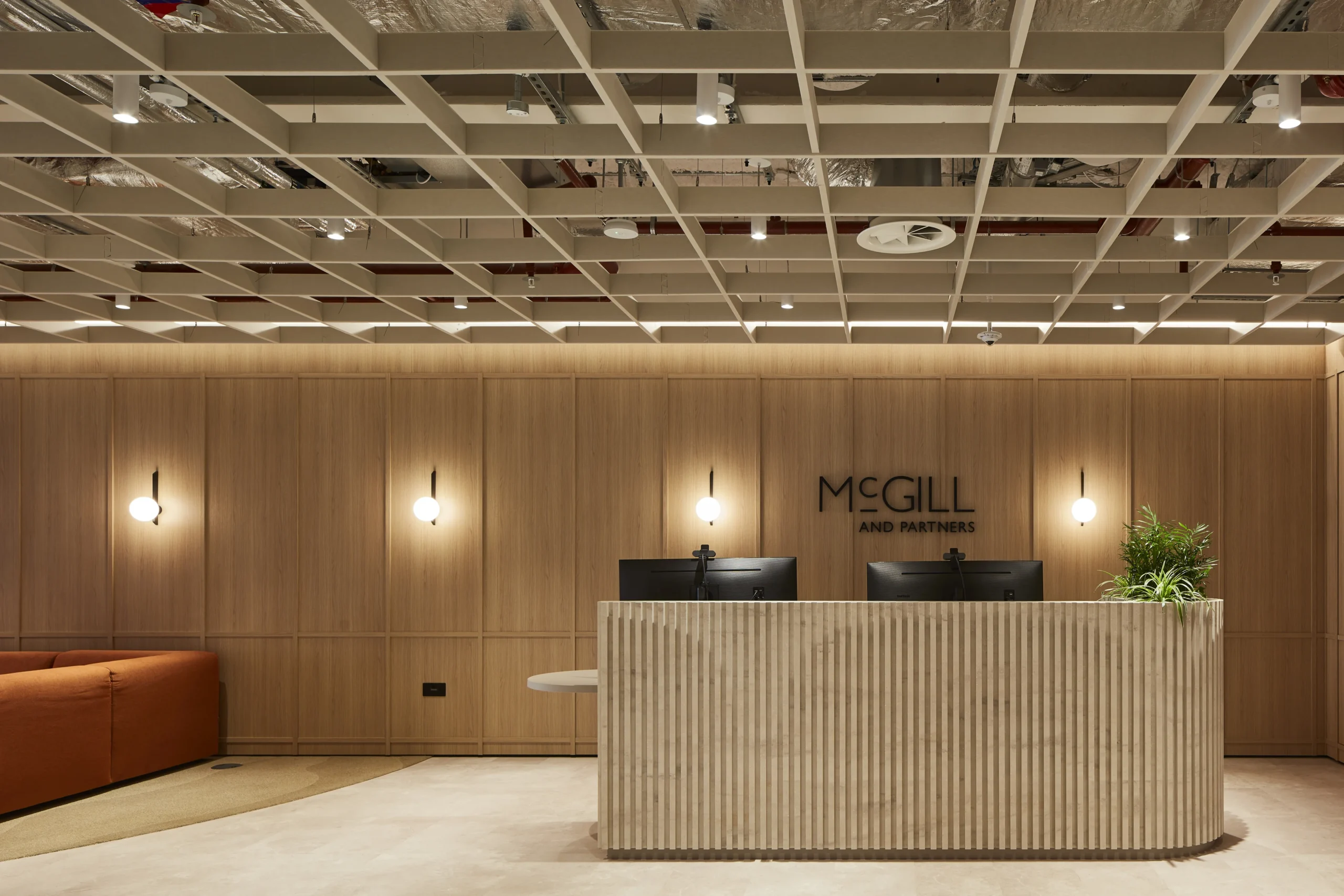 Elegant wooden reception desk with McGill & Partners logo, flanked by stylish pendant lighting and plush seating in a modern office setting.